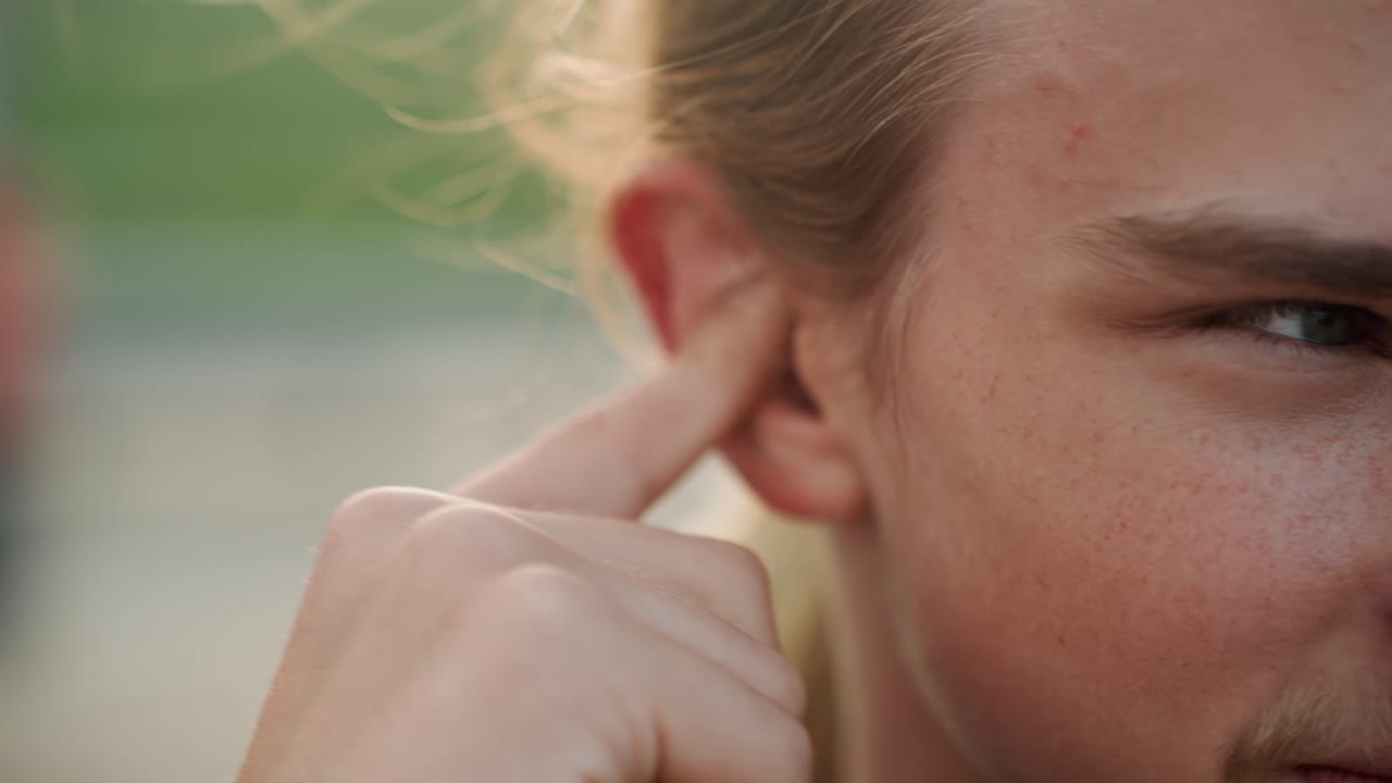 Primo piano di atleta bianco che si aggiusta l'orecchio all'aperto, capelli scompigliati dal vento, dito che preme il canale, regolando l'udito durante l'allenamento. Espressione concentrata con sfondo verde e bagliore del tramonto. Routine sportiva.