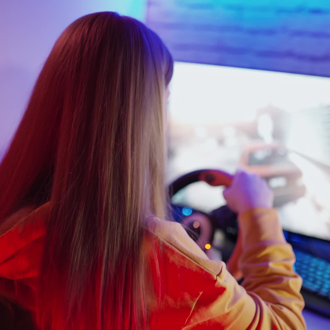 Long-haired girl playing video game. Back view of a young woman with steering wheel playing car racing on a computer at home. Gaming concept.