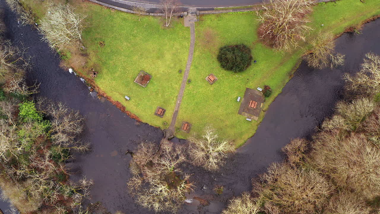 A river winds through a lush green landscape, weaving around dense trees and open grassy areas, drone top down pan up to road