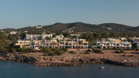 Aerial-forward-view-of-boats-parked-in-a-silent-and-calm-sea-with-a-young-man-standing-at-shore-looking-across-with-house-and-resort-villa-with-greenery-in-Ibiza-in-Spain