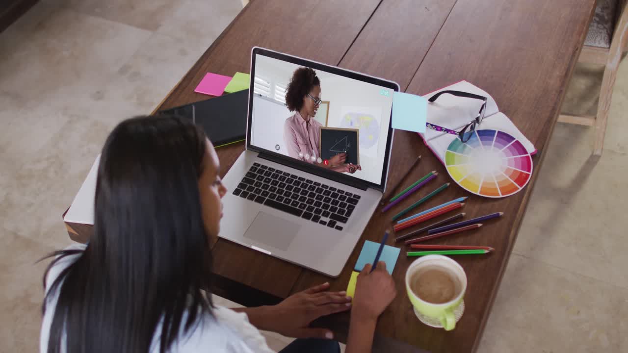 Estudiante caucásica usando una laptop en videollamada con una profesora, tomando apuntes.