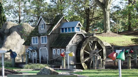 a carefully constructed artful wood house replica sits on the putting green of a golf course as a water wheel turbine spins to and fro into a creek fountain