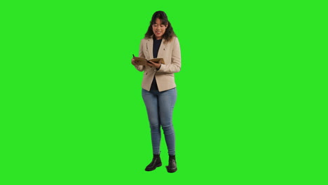 full length studio portrait of female teacher with notebook talking to class standing against green screen 1