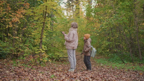 woman in padded jacket writes in notebook while standing on forest trail as girl holds leash of sniffing dog, surrounded by vibrant autumn foliage under calm daylight in quiet woodland setting