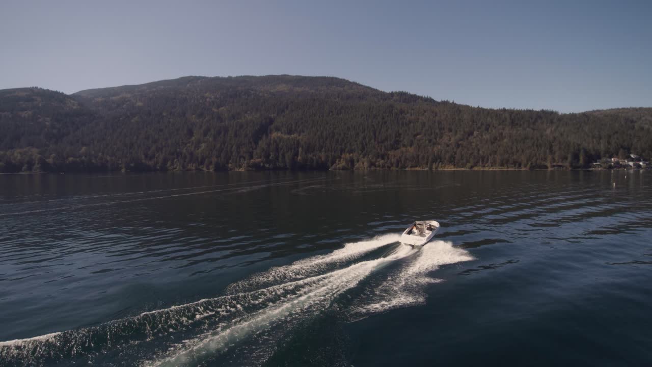 4K-Drohnenaufnahme von Freizeitschnellbooten, die an einem sonnigen Tag auf einem ruhigen Cultus Lake ihre Freizeit verbringen, umgeben von Bergen und Bäumen, mit Wellen auf dem Wasser.