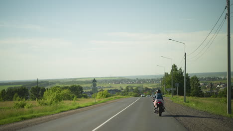 vista trasera de dos amigos montando una bicicleta eléctrica a un ritmo lento en una carretera abierta, los coches son visibles viniendo de la dirección opuesta, con un fondo pintoresco de árboles y una zona residencial distante