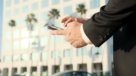 california corporate businessman pulls phone out of pocket to text, palm trees and office building in background