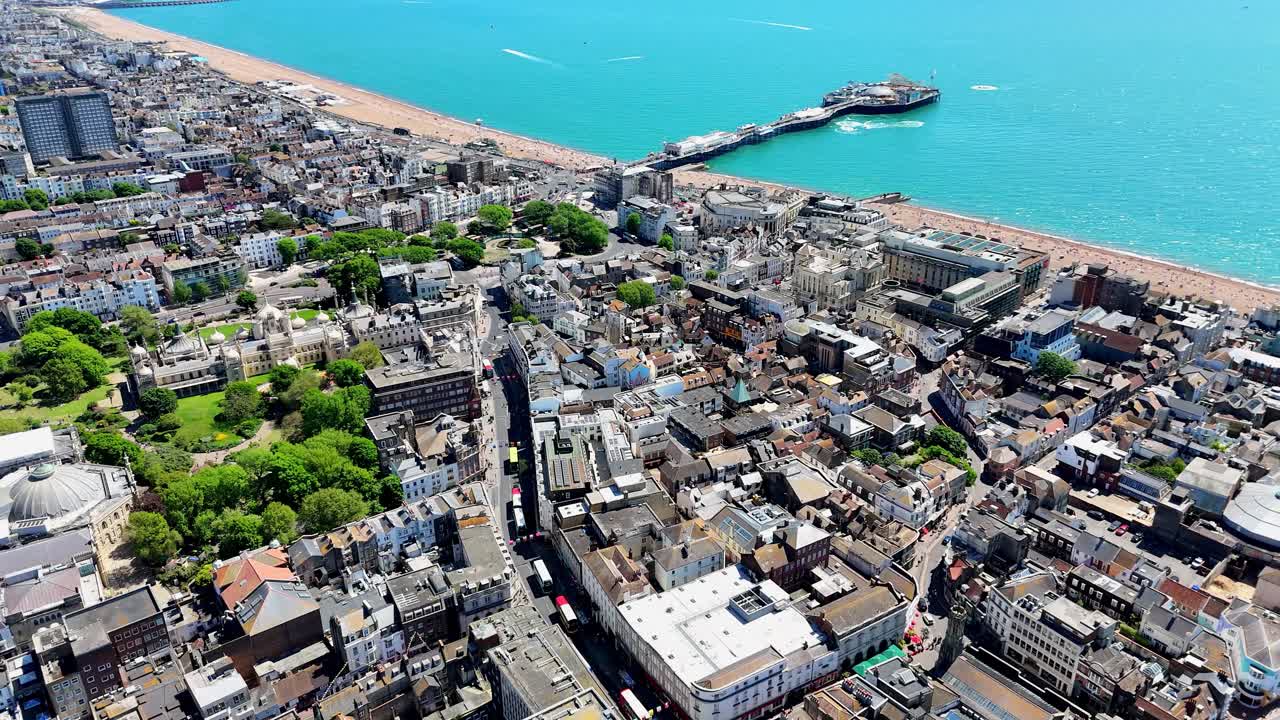 Aerial drone pan over Brighton’s bustling town in summer, featuring the Pavilion, lively streets, and the iconic pier and beach shimmering in the background under sunny skies.