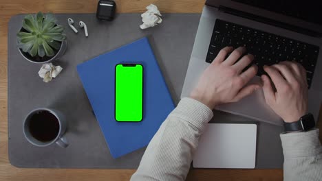 person typing on a laptop with green screen phone on a desk