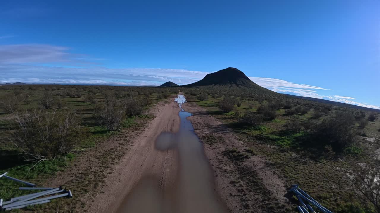 Un drone segue una strada sabbiosa nel deserto con pozzanghere dopo la pioggia in California. Mentre il volo si alza, appare una grande collina isolata, rivelando il vasto paesaggio verde del Mojave sotto un cielo azzurro.