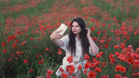 beautiful, cheerful dark-haired girl with a flower in her hair, sitting in a field of wildflowers and red poppies, smiling while taking a selfie with her smartphone
