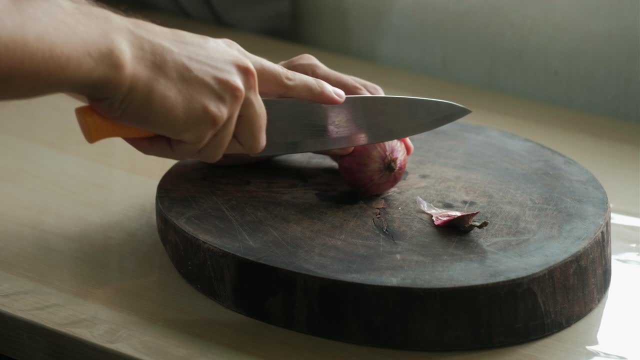 Hands cutting off the root of red onion and cutting in half on wooden chopping board.