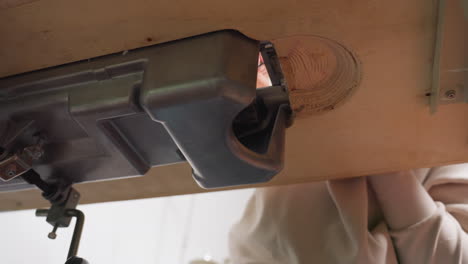 close bottom view of sewing machine installed on wooden table, showing mechanical detail and construction setup, with hands slightly visible underneath as operator works on internal adjustment