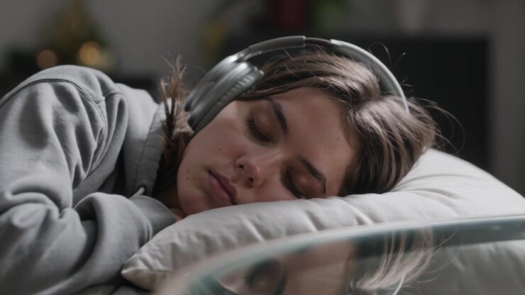 La telecamera si avvicina scorrendo a una donna che dorme sul divano, mettendo a fuoco cuffie argentate, un cuscino e una felpa con cappuccio grigia