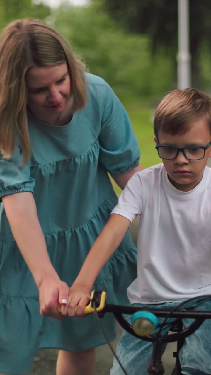 una madre ayuda a su hijo mientras anda infelizmente en bicicleta por una pasarela del parque