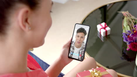 mujer biracial sosteniendo un teléfono inteligente con un hombre biracial en la pantalla con un regalo en el escritorio