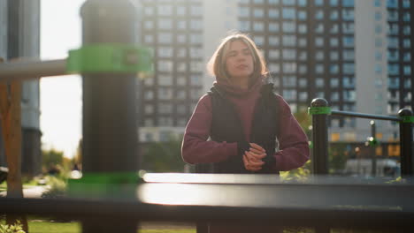 hoodie jacket wearing fitness advocate stands by pullup bars under bright sunlight rolling hands then turning head sideways while gently stretching neck muscles in preparation for outdoor workout