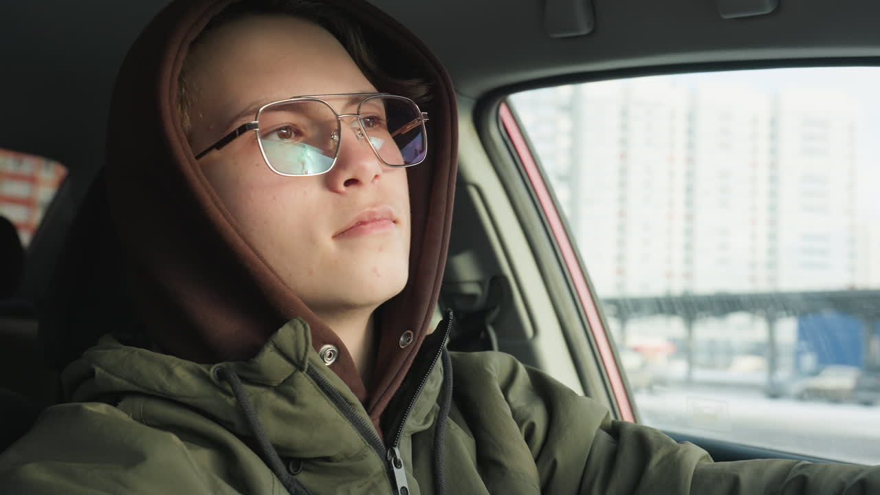 close up of hoodie wearing young man in glasses gazing thoughtfully out car window at urban skyline reflecting inner calm and introspection under soft natural light in parked vehicle
