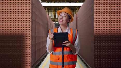 asian female engineer with safety helmet taking note on the tablet and looking around while standing with red brick packed in stacks are stored