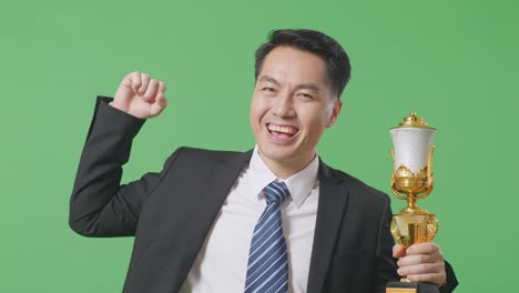 close up of asian business man in a suit and tie with a gold medal and trophy flexing his bicep and smiling to camera on green screen background in the studio