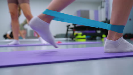 close-up leg view of a person using a resistance band on a yoga mat, another woman walking in blurred background, mirror reflecting the gym surroundings