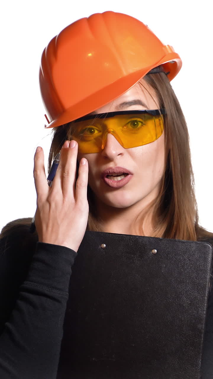 A woman in an orange helmet and protective eyeglasses is talking and holding a pen and folder in her hands on a white background. Features of work of the engineer Vertical video
