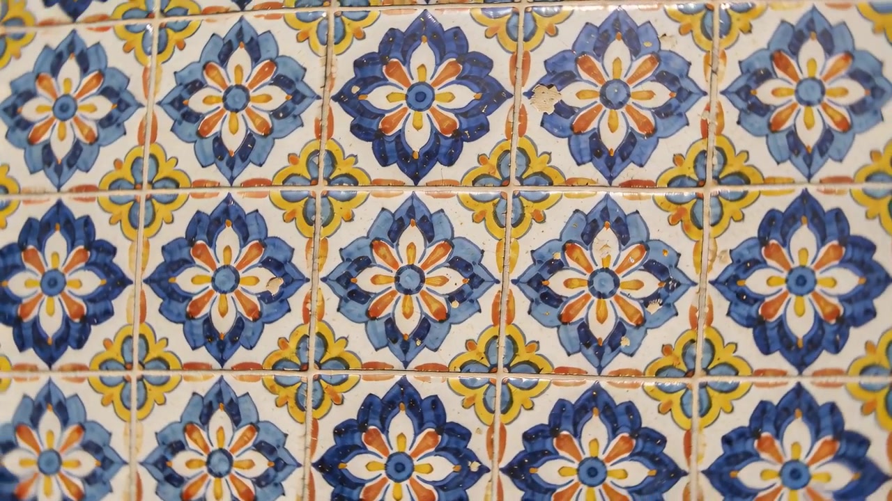 Telecamera in movimento che scorre e ruota su una parete di piastrelle smaltate, rivelando rosette blu, crepe nella malta e lucentezza.