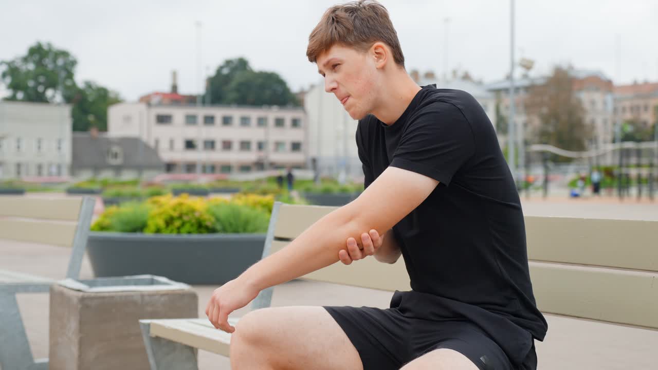 Fixed shot of man in sportswear sitting on bench, bending and extending his elbow while holding it with his other hand