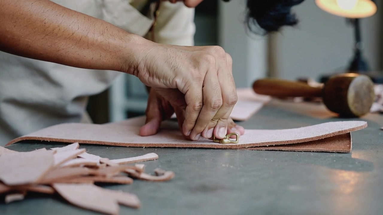 Placing buckle, artisan in apron pressing it into leather panel at bench securing leather piece