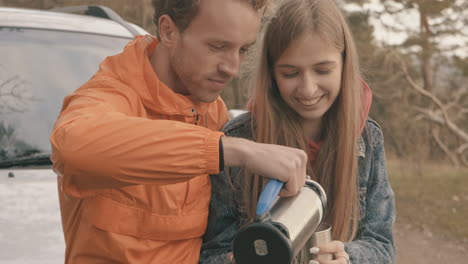 young couple sitting on the car having a hot drink with a thermos