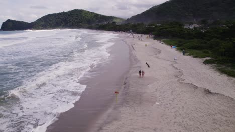 drone volando sobre la orilla mientras la gente camina cerca de las olas