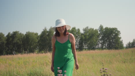 woman bending forward to smell lavender flowers in tall grass meadow wearing green dress and white hat under clear blue sky with distant treeline and breeze stirring blades