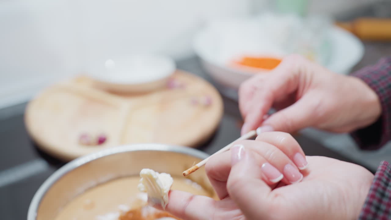 vista de cerca de la mano de una persona que sostiene mantequilla cremosa en la punta del dedo, preparándose para dejarla caer en una olla de papel marrón liso en una cocina brillante