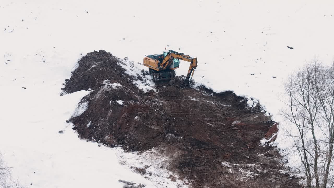 Drone video captures powerful excavator clearing snow-covered land in winter landscape. Prospective, frozen hills, earth-moving machinery amidst the stark contrast of white snow