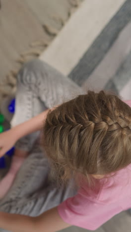 little girl with french braid sits on rug building wall from constructor. preschooler plays with small multi-colored blocks upper close view