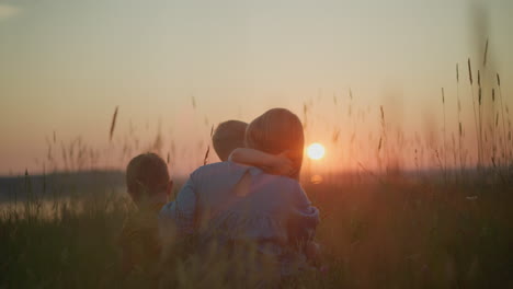 a back shot of a mother in a blue gown sitting in a field during sunset, holding her younger child on her lap while the older child sits beside her. they all gaze out at the serene lake