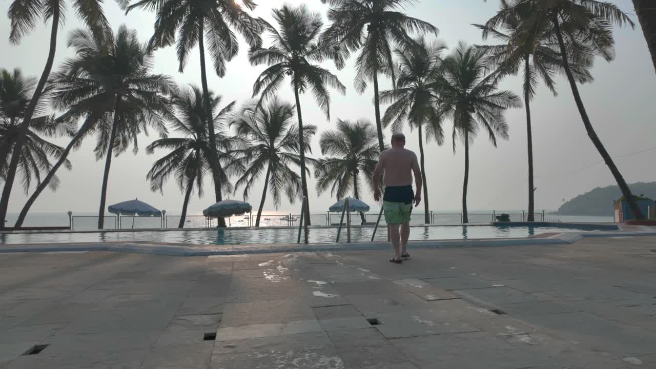 4K video of an older man going to swim in a luxury resort's pool surrounded by palm trees in Goa, India