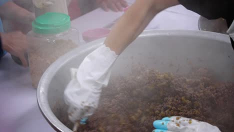 Woman-placing-powder-flavoring-into-a-large-bowl-of-purple-rice-and-pork-meat-filling-being-stirred-and-mixed-by-another-person