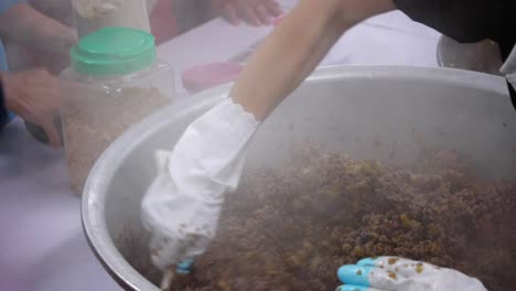 mujer colocando saborizante en polvo en un tazón grande de arroz morado y relleno de carne de cerdo que otra persona agita y mezcla