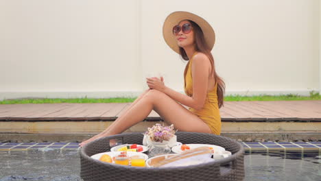 a young woman in a bathing suit sitting on the edge of a pool reaches down to steady the floating breakfast platter
