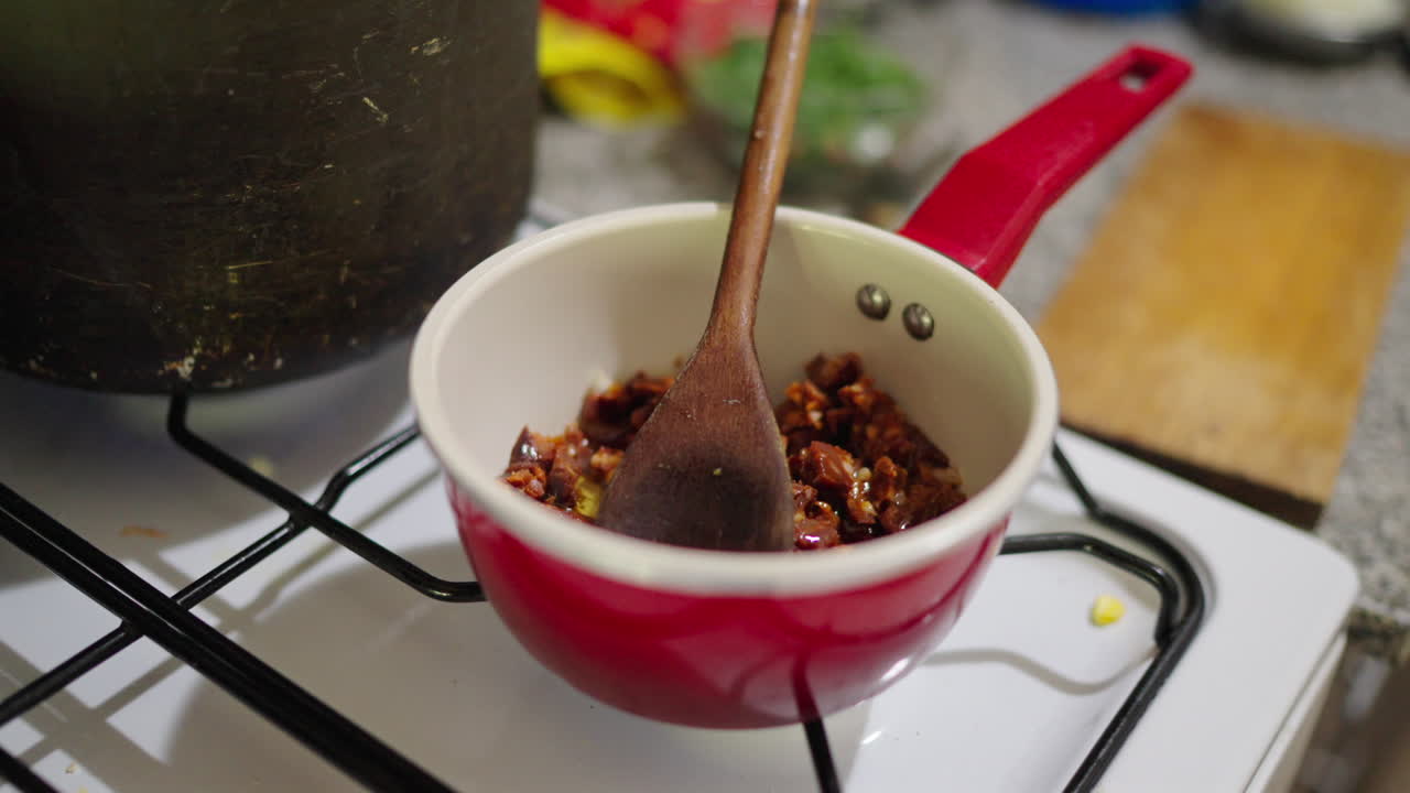Herbs and spices fry in red sauce for Argentinian locro, seen in small metal pan, hands pour in oil