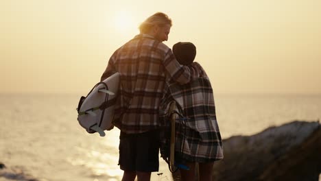 Rear-view-of-a-happy-couple,-a-blond-guy-with-a-beard-in-a-checkered-shirt-stands-with-his-blonde-girlfriend-in-a-black-hat-and-a-checkered-shirt,-they-hold-surfboards-in-their-hands-and-look-towards-the-sea-in-the-morning-at-sunrise