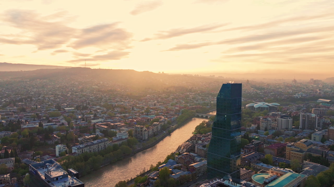 Drone shot over Tbilisi, Georgia during sunrise, capturing warm golden hour tones across the river, historic districts, and surrounding hills from above.