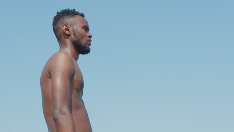 a black man with a beard in bare torso looking around against a bright blue sky