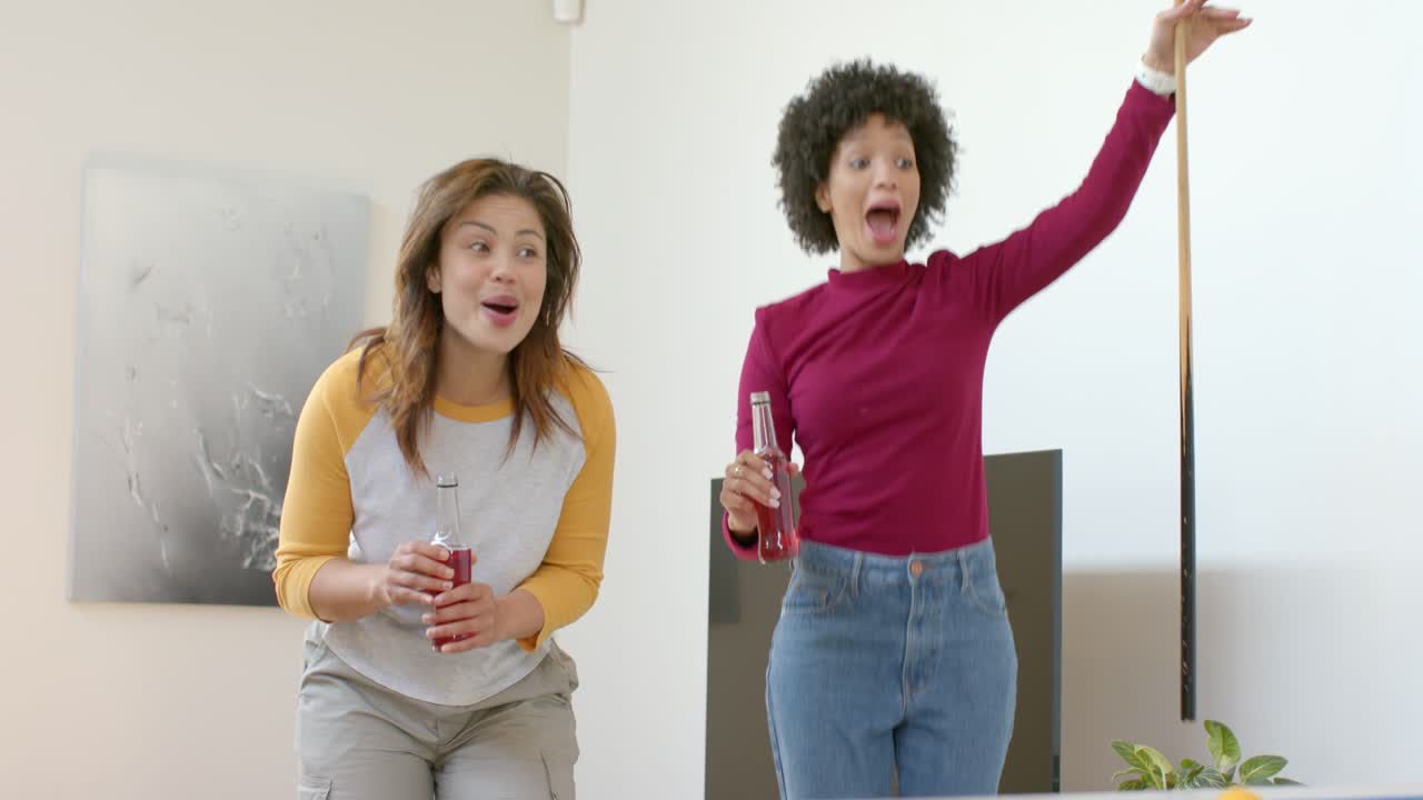 Diverse female friends celebrating shot after break shot scattering balls on pool table at home