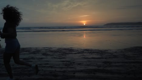 Vista-Panorámica-En-Cámara-Lenta-De-Una-Silueta-De-Mujer-Joven-Corriendo-En-Una-Playa-De-Arena-Junto-Al-Océano,-Al-Amanecer