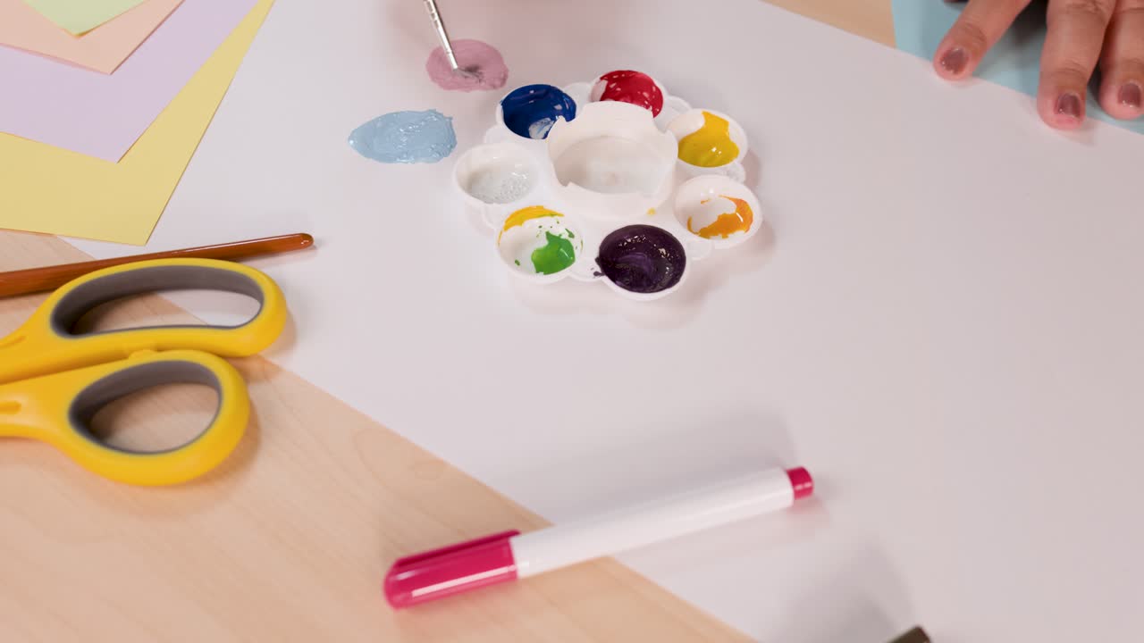 Person mixes watercolor paints on palette, painting on white paper in bright, well-lit workspace