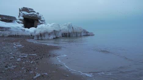 small waves breaking against the ruins of karosta northern forts fortification on the shore of baltic sea on a cloudy winter day, covered with ice, snow and icicles, wide shot