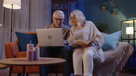 senior grandparents couple reading book, using laptop pc on couch in night living room at home