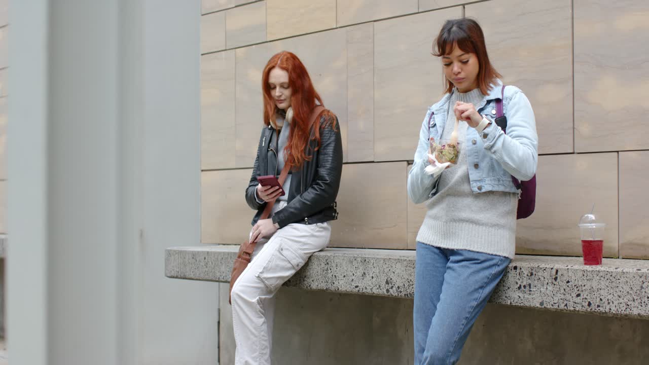 Подруги обедают на скамейке возле офиса, просматривают ленту в смартфоне и едят салат